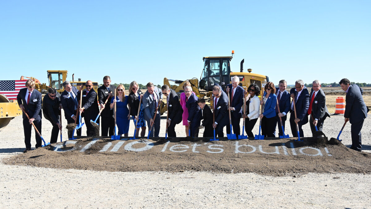 A group of people standing in a line holding shovels at a groundbreaking ceremony. Behind them are construction vehicles and a pile of dirt with the words lets build! written on it. The sky is clear and blue.