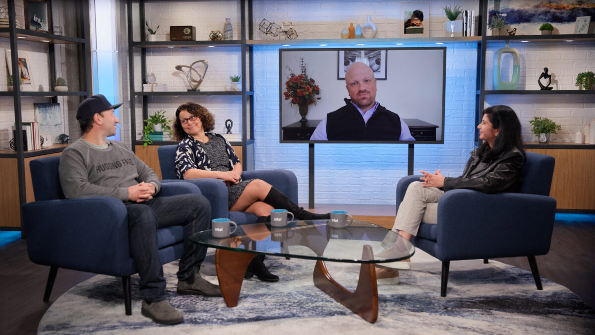 Four people are participating in a video conference; three sit in blue armchairs while one person appears on a large screen. The room is modern, with decorative shelves and soft lighting. A coffee table with mugs and a rug are in the foreground.