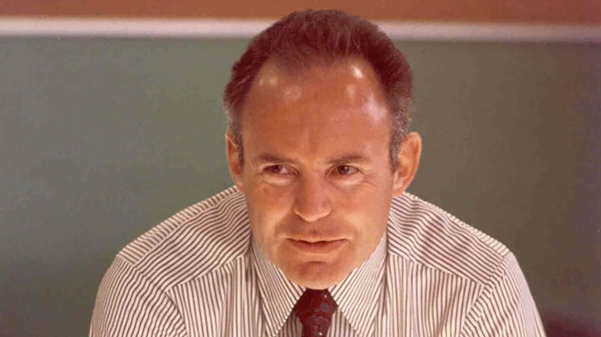 A man with short hair is wearing a striped shirt and tie. He is looking slightly to the side with a neutral expression. The background is a blurred wall with soft colors.