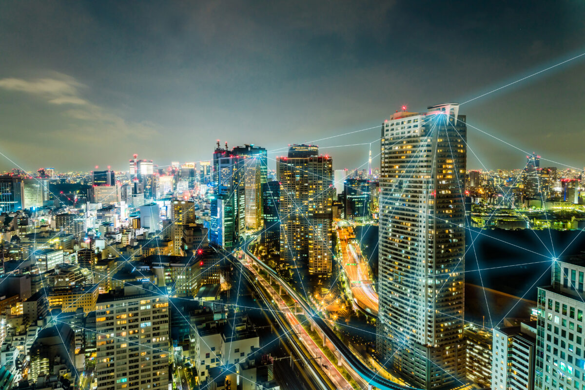 A vibrant cityscape at night features tall skyscrapers with illuminated windows. Light trails from vehicles show traffic on the roads. Digital network lines are superimposed over the image, suggesting connectivity or communication networks.