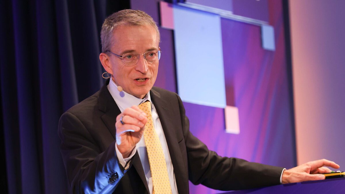 A man in a suit and tie is speaking on stage, holding a small object in his hand. He wears glasses and has a microphone attached to his face. The background is a mix of purple and pink hues with geometric designs.