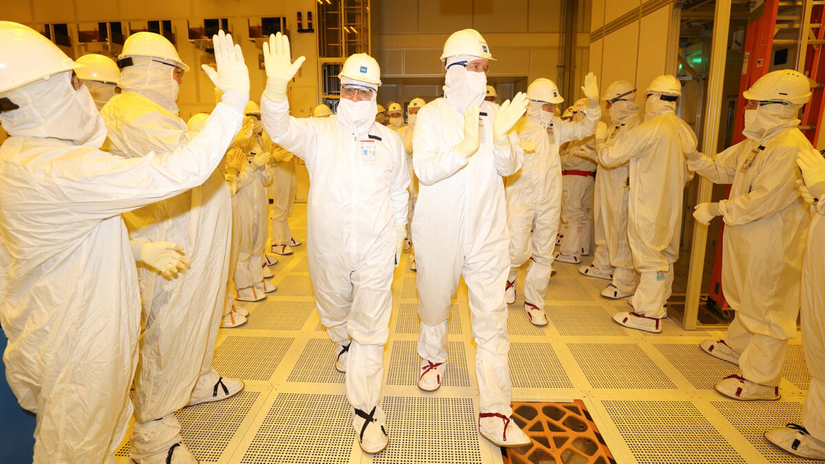 A group of people wearing white cleanroom suits and helmets gather in a brightly lit industrial facility. Two individuals in the center gesture with their hands while others observe or gesture back. The setting appears to be a high-tech environment.