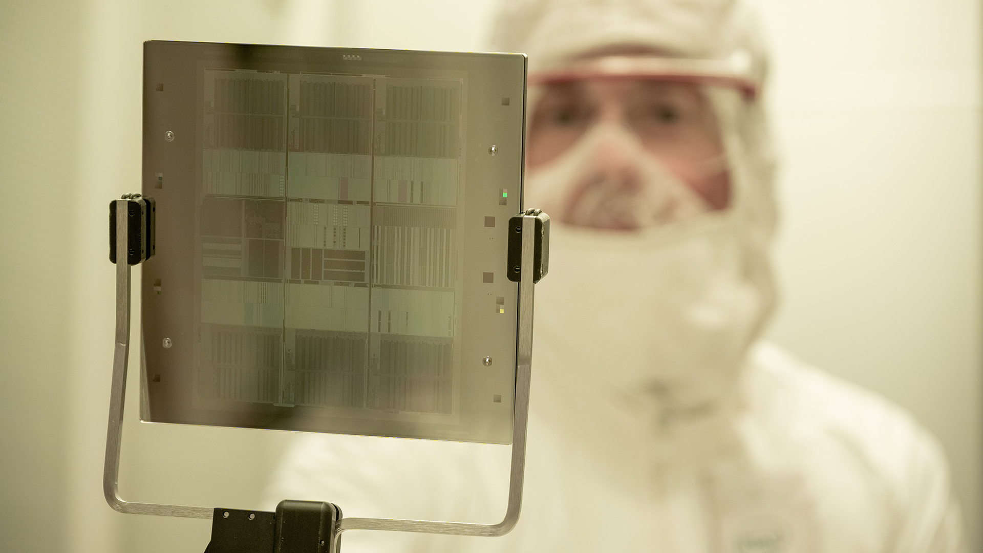 A person in a cleanroom suit with a face mask and protective goggles holds a semiconductor wafer. The background is blurred, emphasizing the wafers intricate patterns.