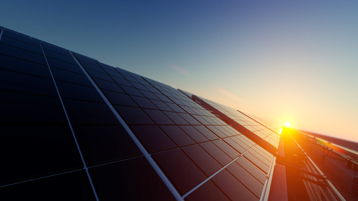 Close-up of solar panels with a sunrise in the background. The panels are angled towards the sun, capturing its early morning light. The sky is clear, with warm hues of orange and blue blending on the horizon.