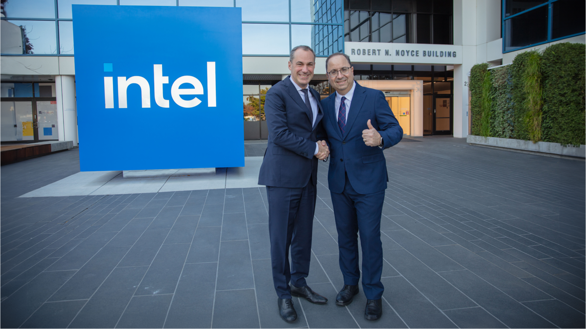 Two businessmen shaking hands and smiling in front of the Intel sign at the Robert N. Noyce Building. One man is giving a thumbs up. The background shows modern architecture and greenery.