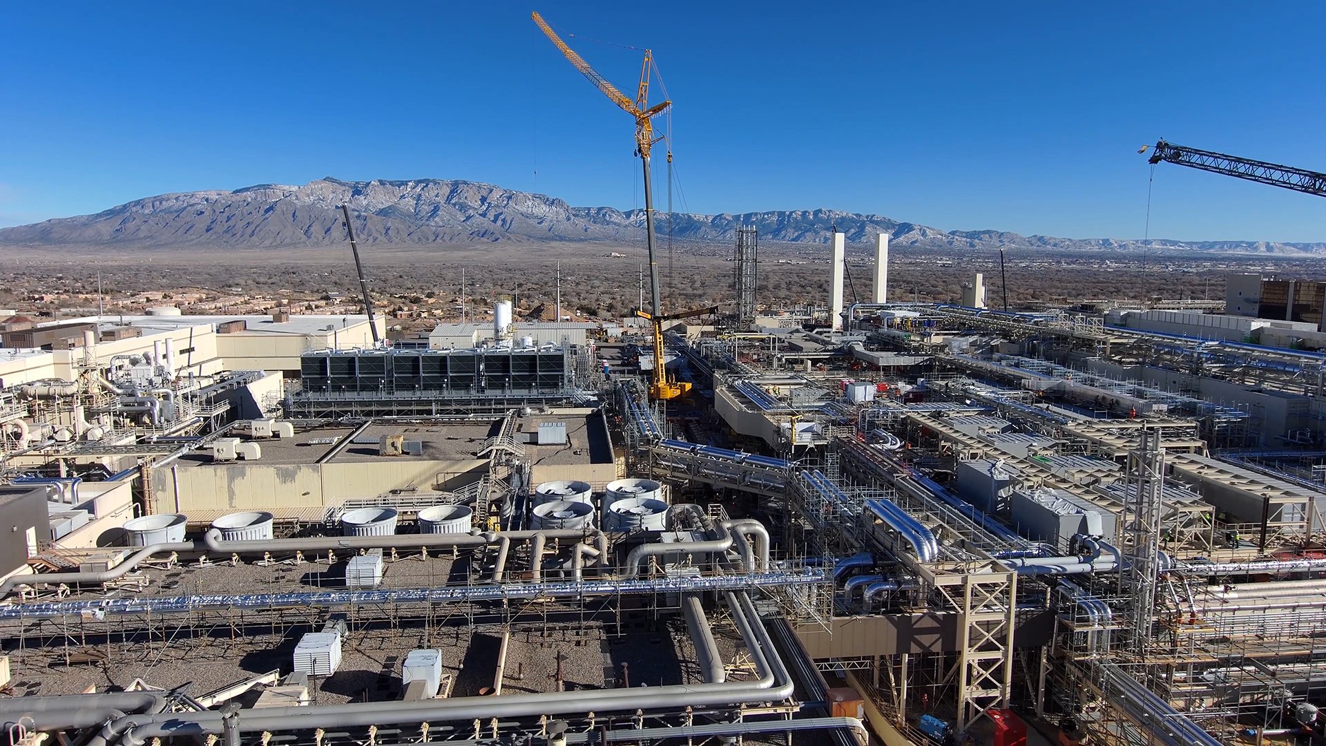 A large industrial complex with numerous pipes, buildings, and cranes under a clear blue sky. In the background, there are mountains and a barren landscape. The scene suggests an active construction or industrial site in a desert area.