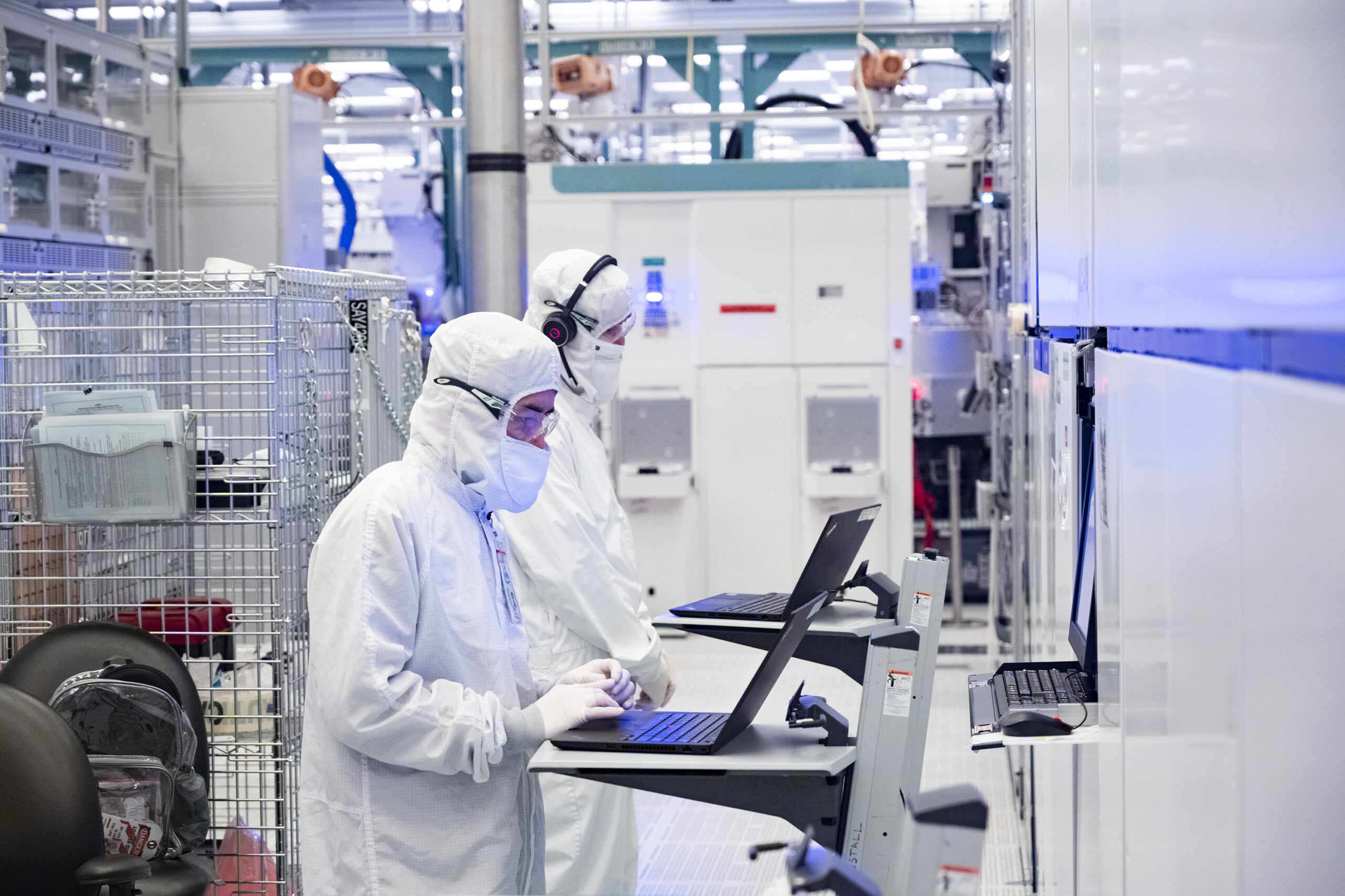 Two workers in full-body protective suits and masks are operating laptops in a cleanroom environment, likely a semiconductor manufacturing facility. The area is filled with high-tech equipment and bright lighting.