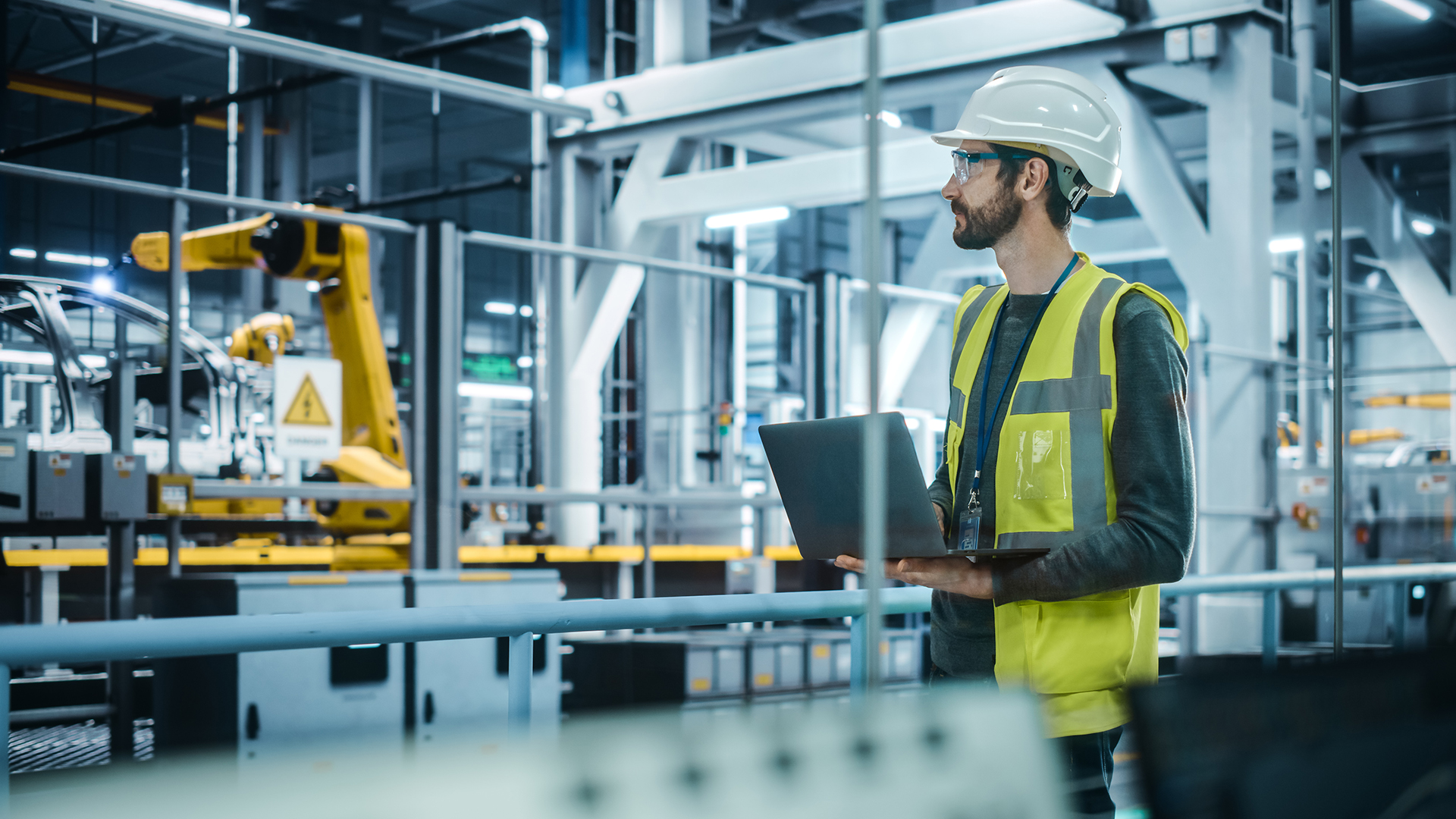 A man in a hard hat and safety vest holds a laptop in an industrial setting. He stands in front of machinery and robotic equipment, appearing focused on his tasks. The environment is well-lit with metal structures.