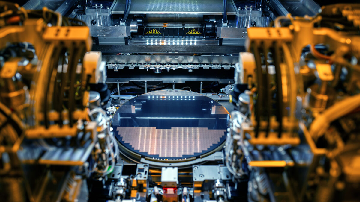 A high-tech machine processes a silicon wafer in a cleanroom environment, showcasing intricate components and precise engineering. The setting is brightly lit, emphasizing the delicate nature of semiconductor manufacturing equipment.