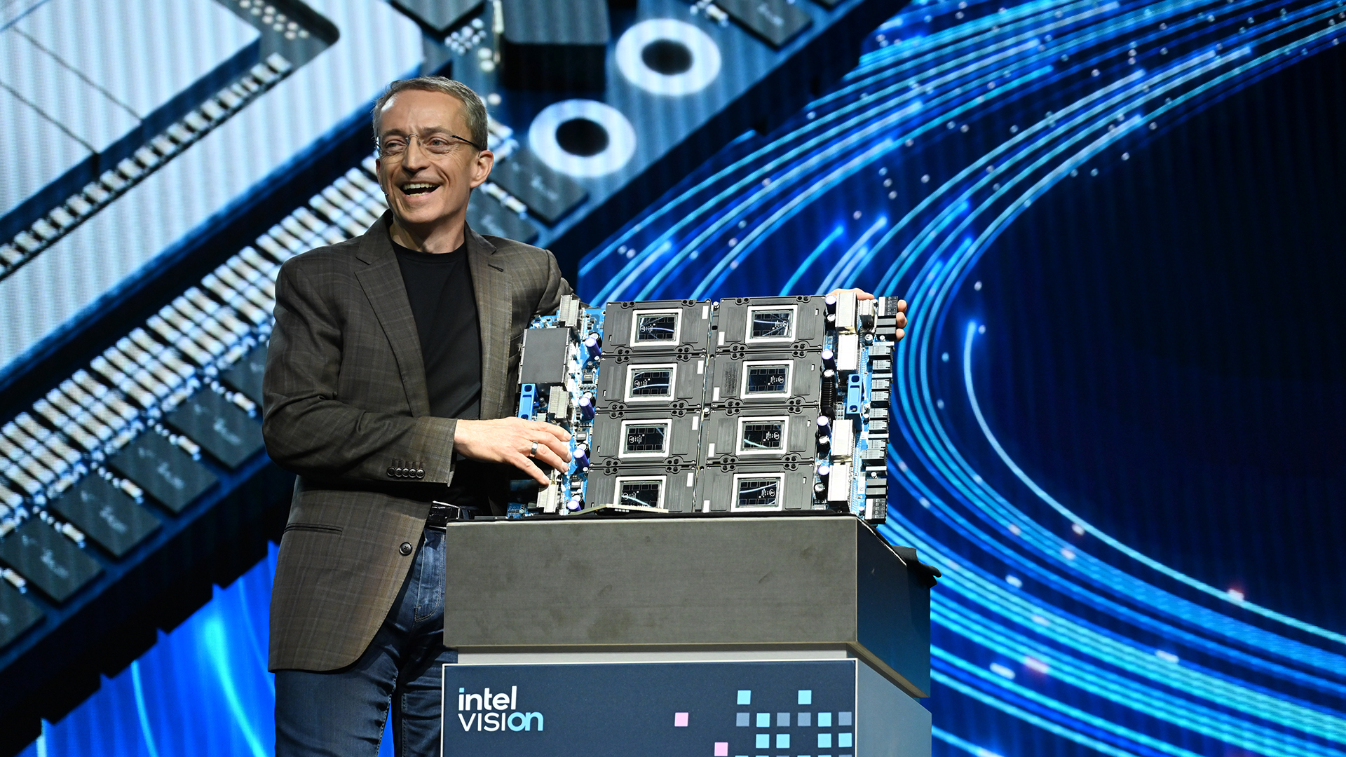 A person in a suit stands on stage, smiling, and holding a large piece of computer hardware. The stage backdrop is a graphic of circuit patterns and digital designs. A sign reads intel vision.