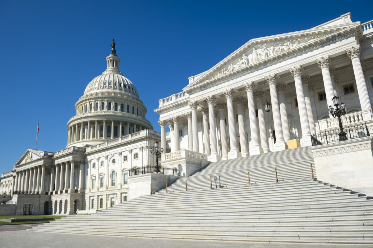 The image shows the United States Capitol building in Washington, D.C., featuring its iconic dome and neoclassical style architecture. A clear blue sky forms the backdrop, and wide steps lead up to the entrance.