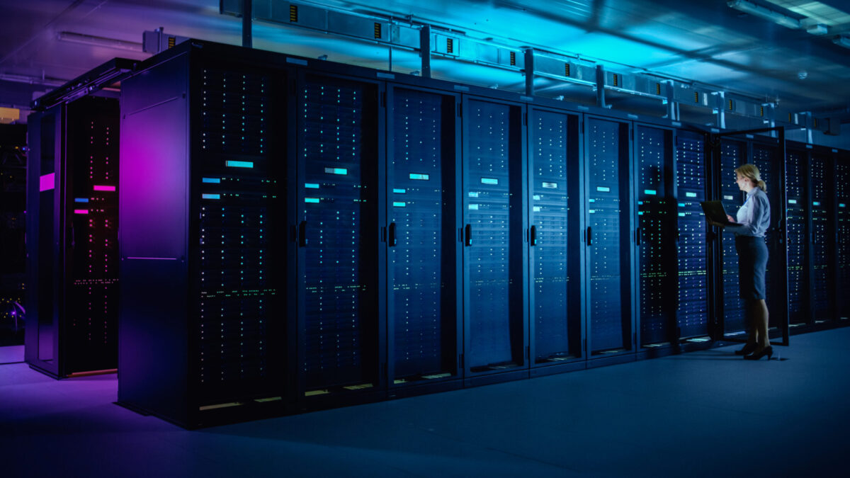 A person stands in a data center, working on a laptop next to a row of tall, illuminated server racks. The room is bathed in blue and purple lighting, creating a modern and high-tech atmosphere.