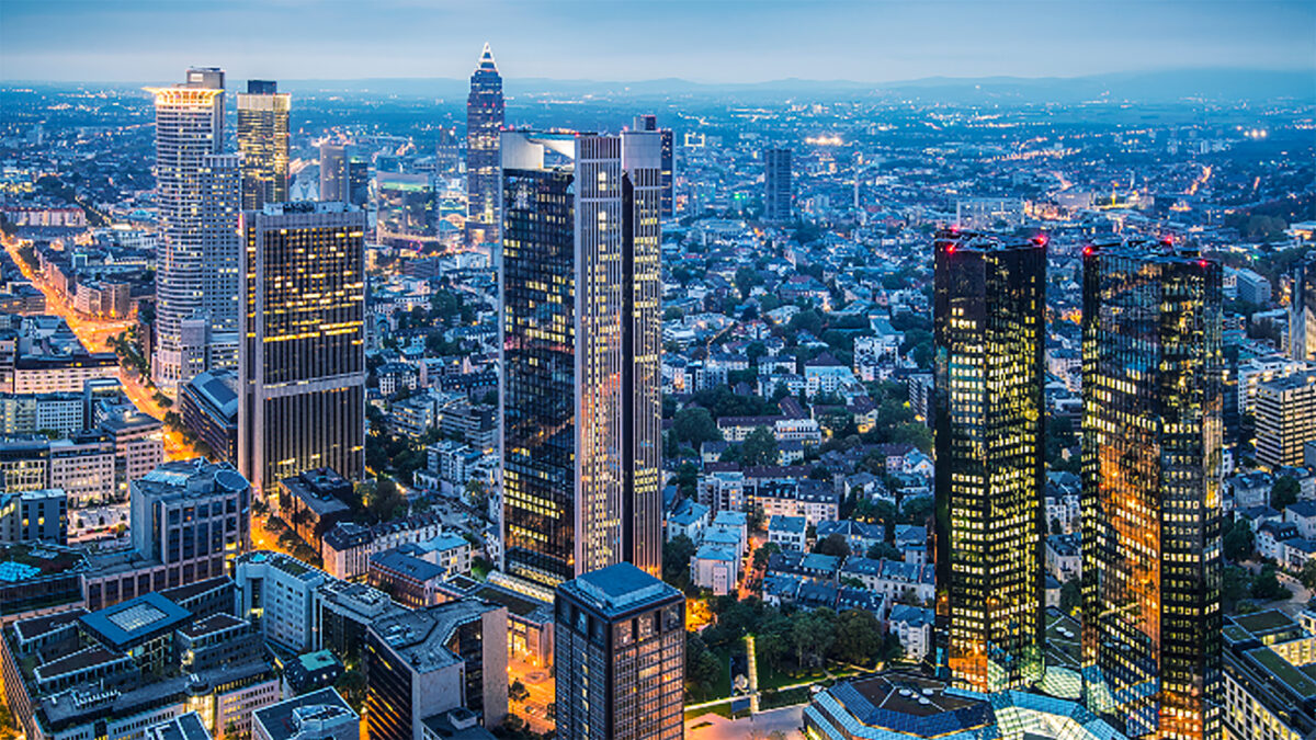 Aerial view of a cityscape at twilight featuring tall, illuminated skyscrapers and a mix of modern and older buildings. Streets are lined with lights, and the sky is a deep blue, indicating early evening. Hills are faintly visible in the background.