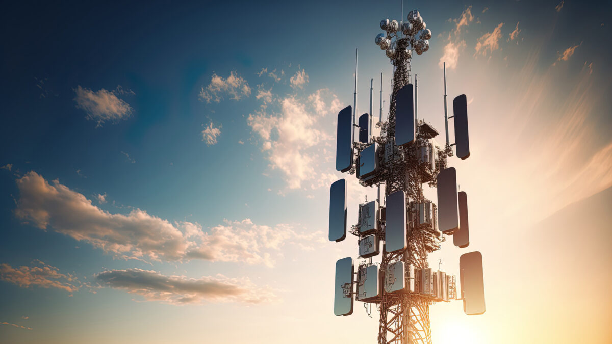 A tall communication tower with multiple antennas and dishes stands against a backdrop of a blue sky with scattered clouds. The sun is setting, casting warm light and creating silhouettes of the towers structures.