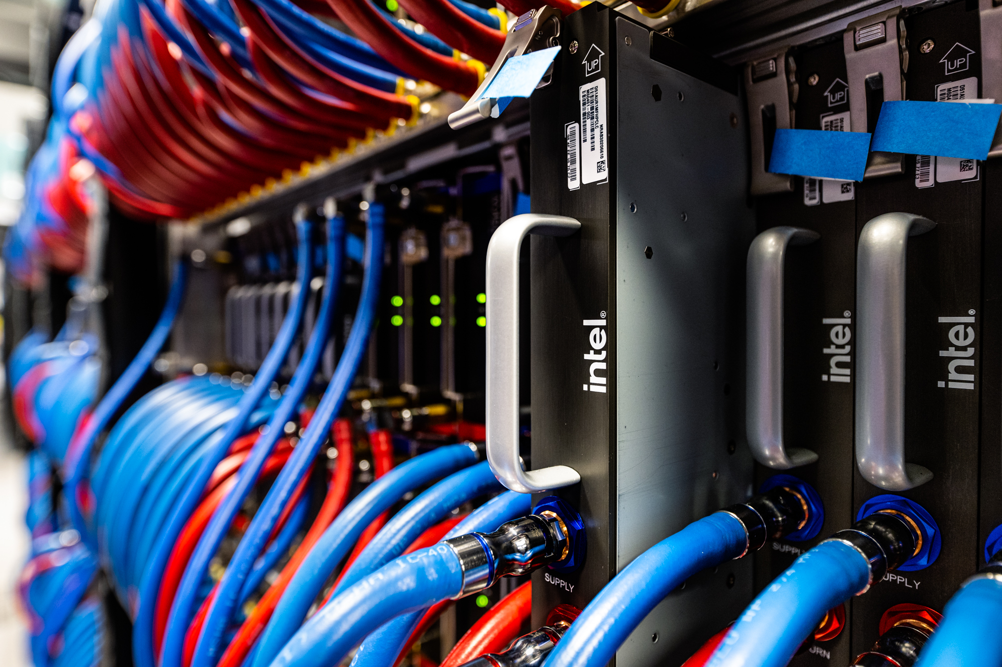 Close-up of server racks with cables and Intel components. Blue and red cables are organized and connected to devices, with green lights and labels visible. The focus is on the hardware connections within a data center environment.