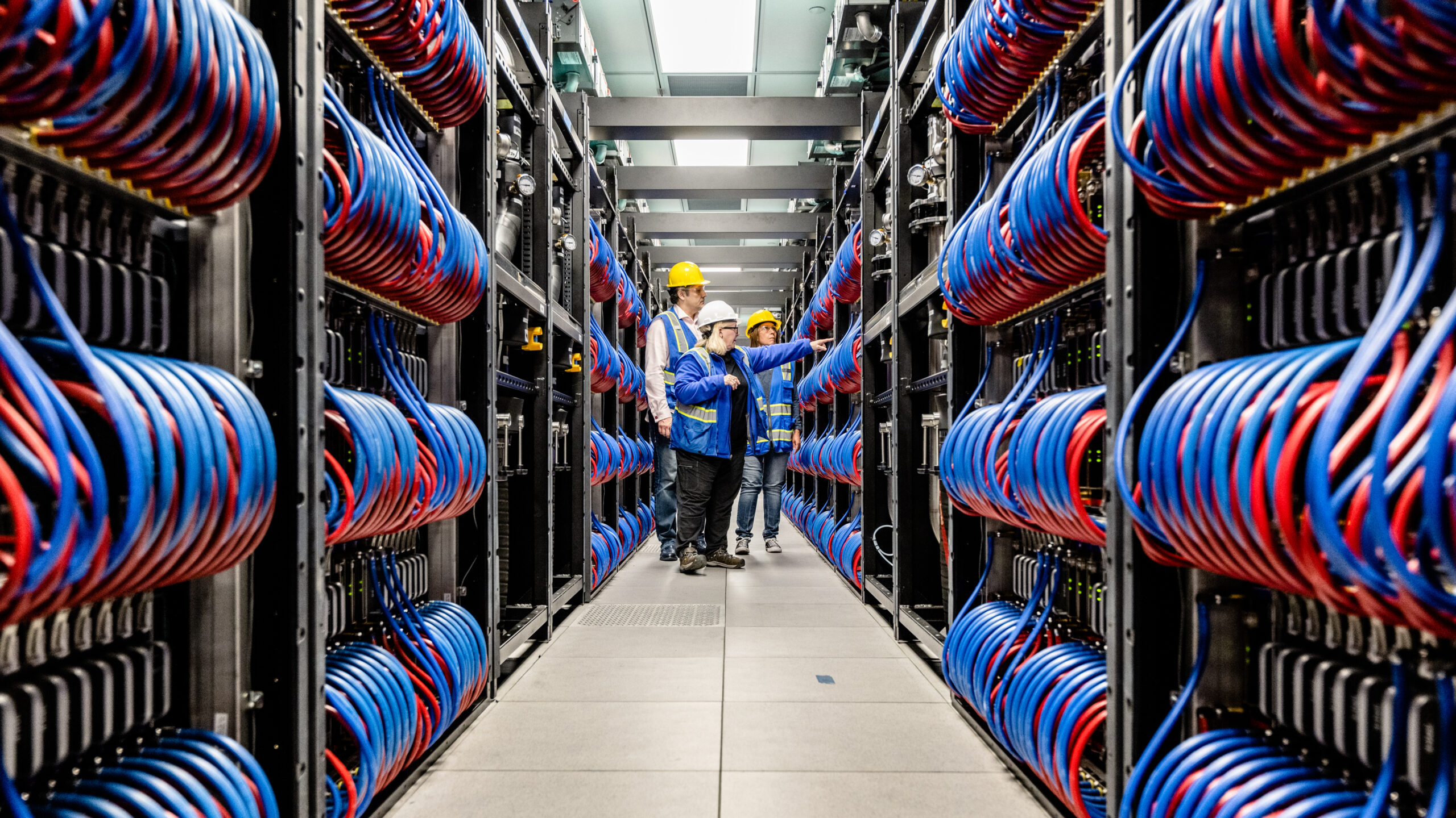 Three technicians in safety gear examine rows of servers with red and blue cables in a large data center. Bright lighting highlights the organized, high-tech environment.