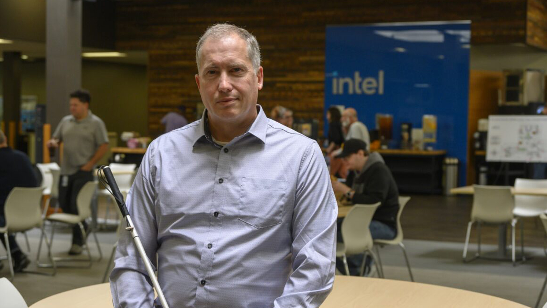 A man in a light gray shirt stands indoors holding a cane, with a blurred background featuring a blue Intel logo. There are several people seated at tables, engaged in various activities.