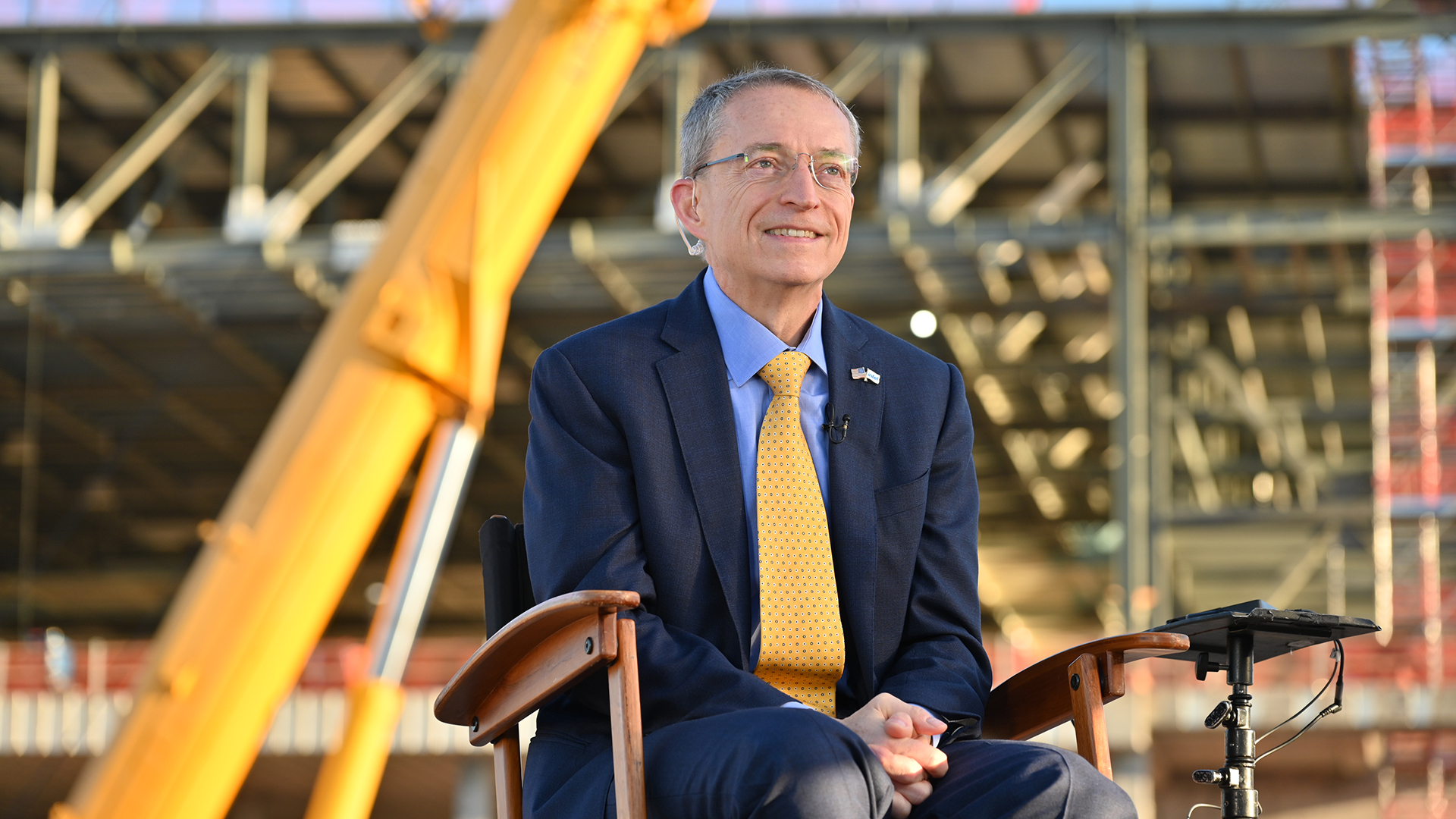 A man in a blue suit and yellow tie sits on a chair outdoors at a construction site. Behind him, a large crane and partially built structure are visible. He appears to be speaking or giving an interview.