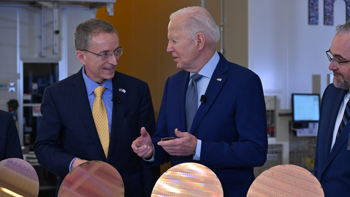 Three men in business suits are engaged in conversation at an event, standing behind a series of semiconductor wafers. They appear to be in a technological or industrial setting.