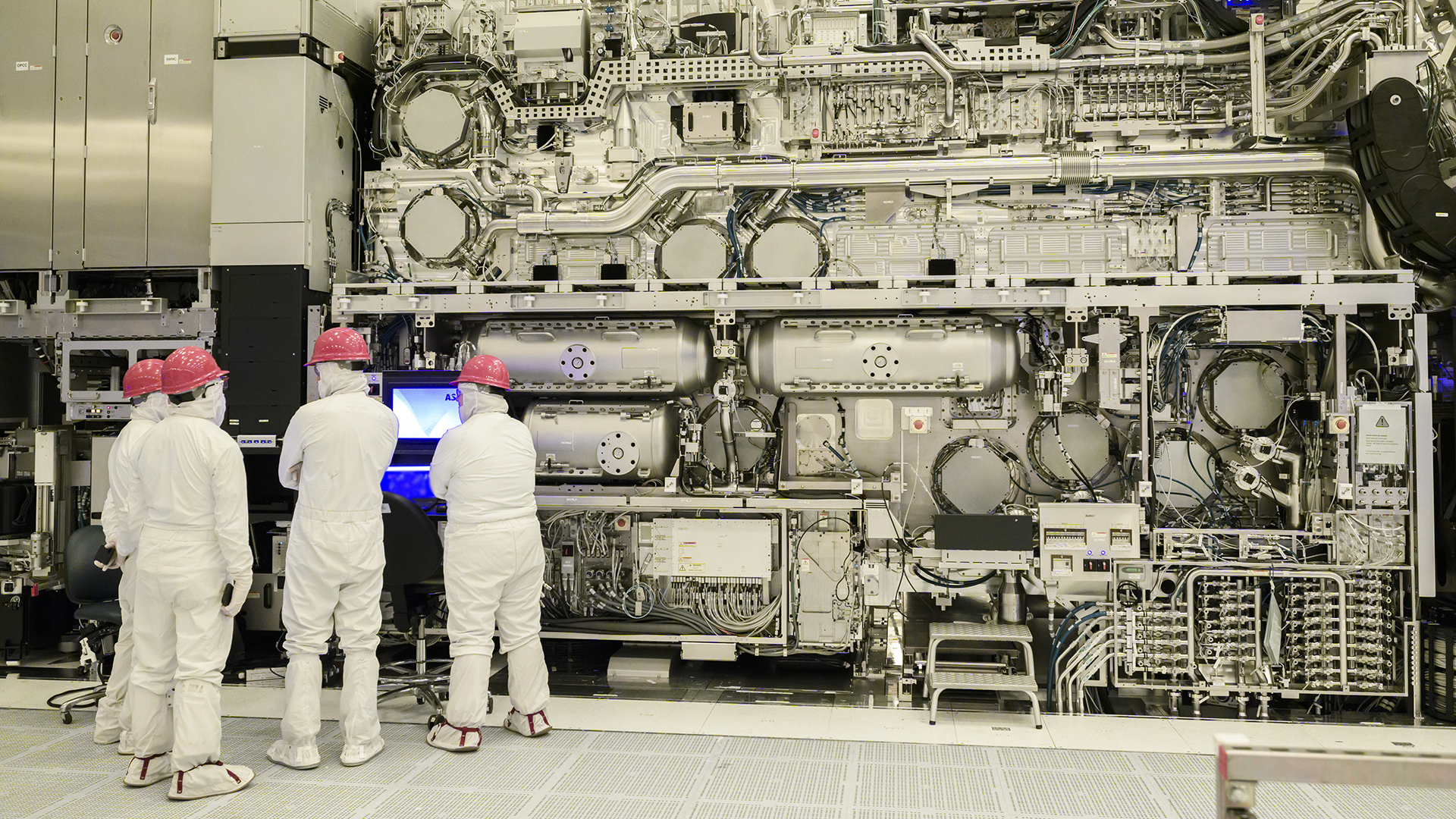 Technicians in protective clothing and pink helmets work on a large, complex machinery in a high-tech industrial setting, facing a wall of intricate pipes and components, with one operating a computer screen.