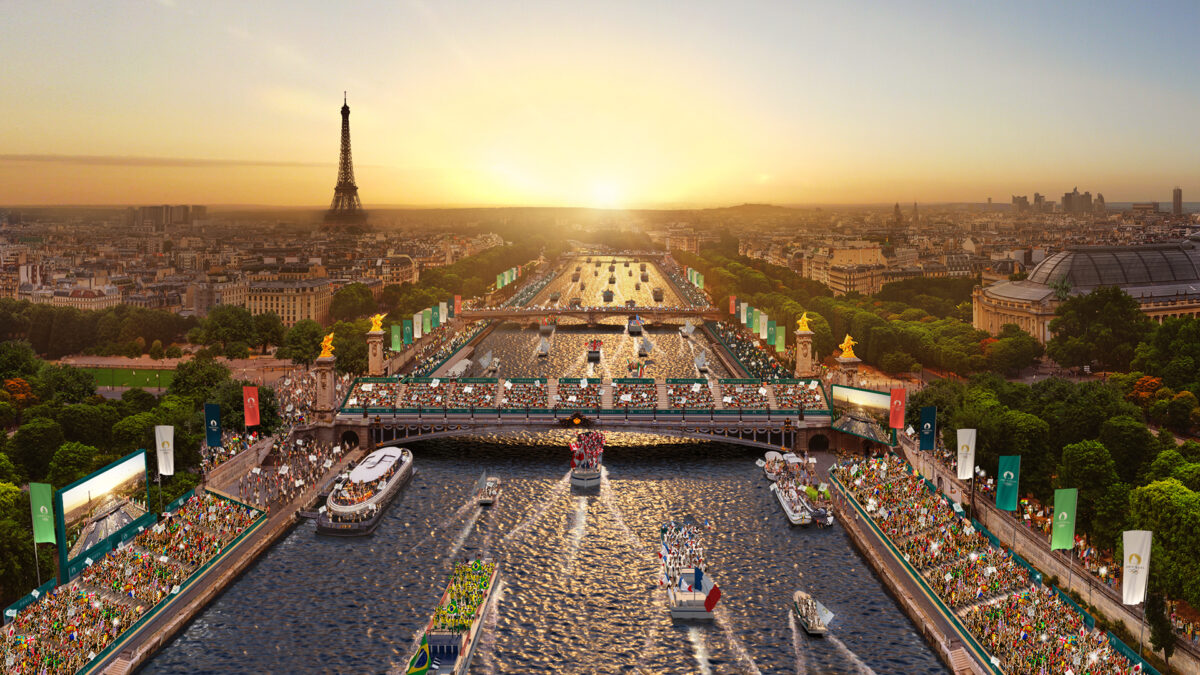 Aerial view of a river parade in Paris during sunset, with the Eiffel Tower in the distance. Boats move along the river, and large crowds gather on either side. The cityscape and lush green trees are visible under a golden sky.