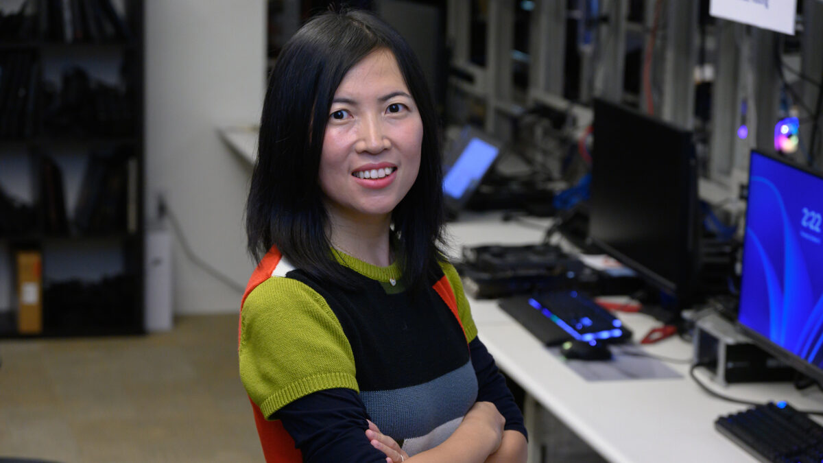 A woman with long black hair smiling and standing with folded arms in an office environment. She is wearing a multicolored sweater. Behind her are desks with computers and shelves with equipment.