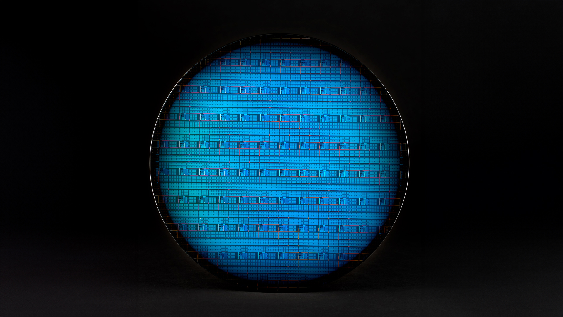 A close-up view of a silicon wafer with intricate blue circuitry patterns against a dark background. The circular wafer showcases a grid-like design, emphasizing the precision and complexity of microchip technology.