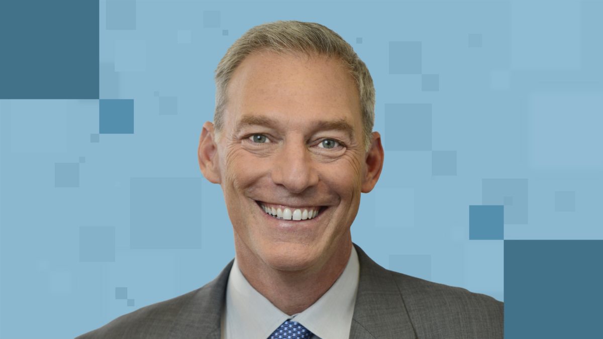 A person with short, gray hair smiles warmly in a professional headshot. They are wearing a dark suit and a blue tie. The background features a digital, geometric pattern in shades of blue.