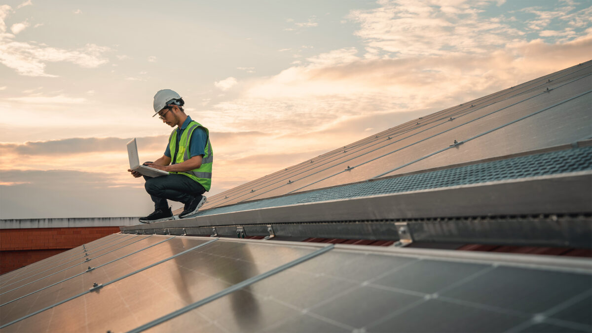 A technician wearing a hard hat and safety vest sits on a solar panel roof, working on a laptop. The sky is partly cloudy with a warm sunset glow.