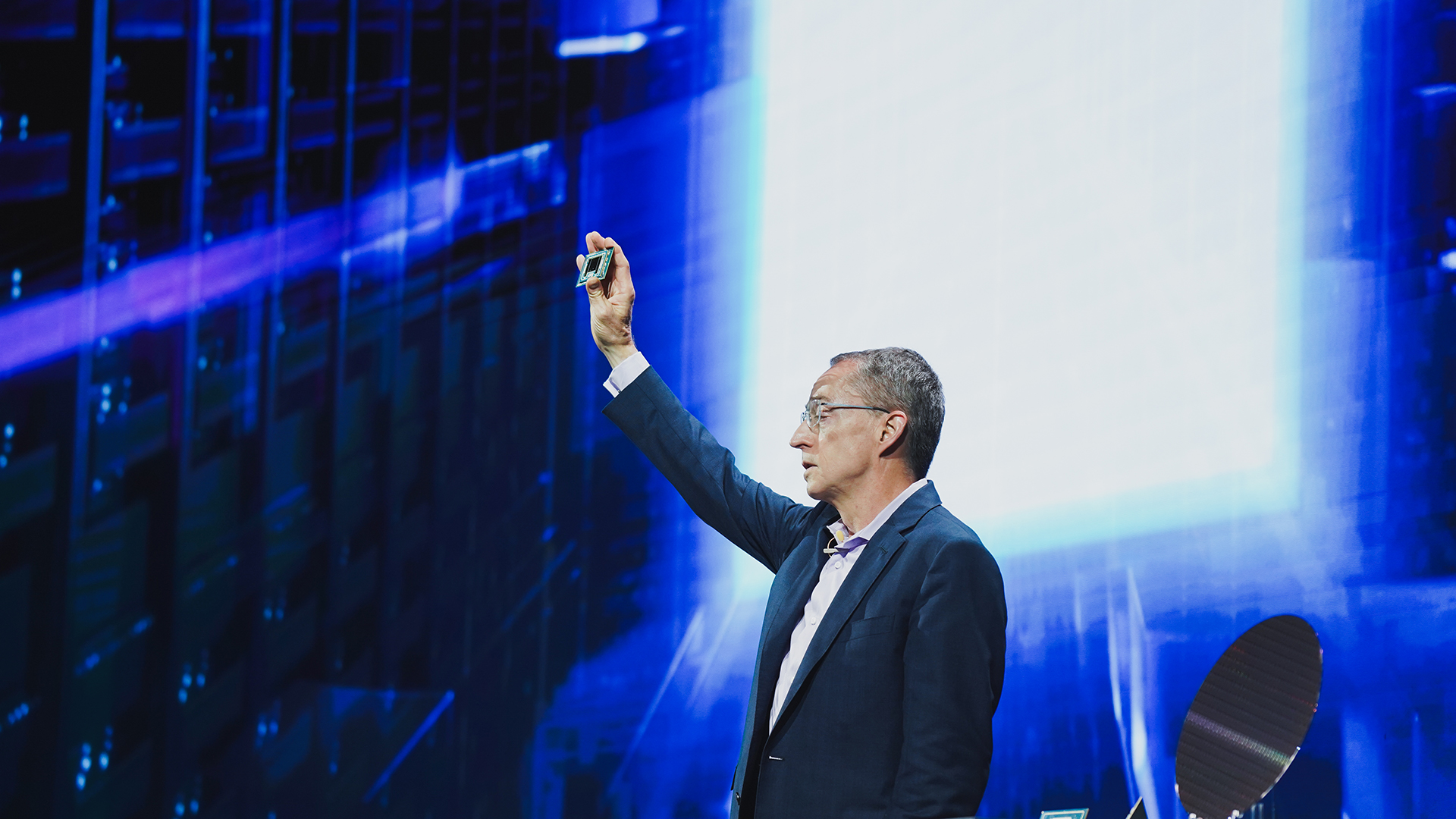 A man in a suit holds up a small electronic component on stage, with a futuristic blue and white background. The scene suggests a technology presentation or conference.