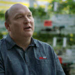 A person in a blue shirt with a red logo sits in a greenhouse filled with plants. The background is blurred and sunlit, highlighting rows of greenery.