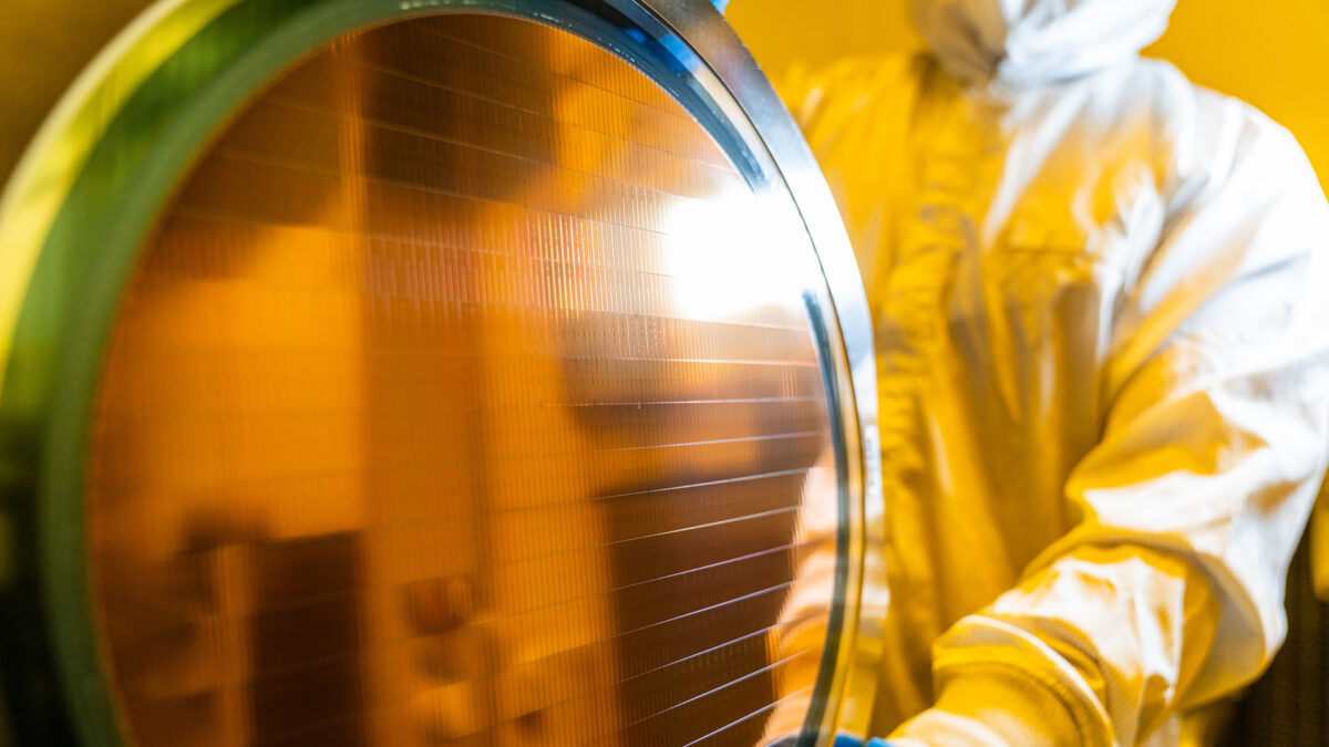 A person in protective gear inspects a large wafer in a cleanroom environment. The wafer reflects the light and has a copper-toned surface. The setting is a semiconductor manufacturing facility.