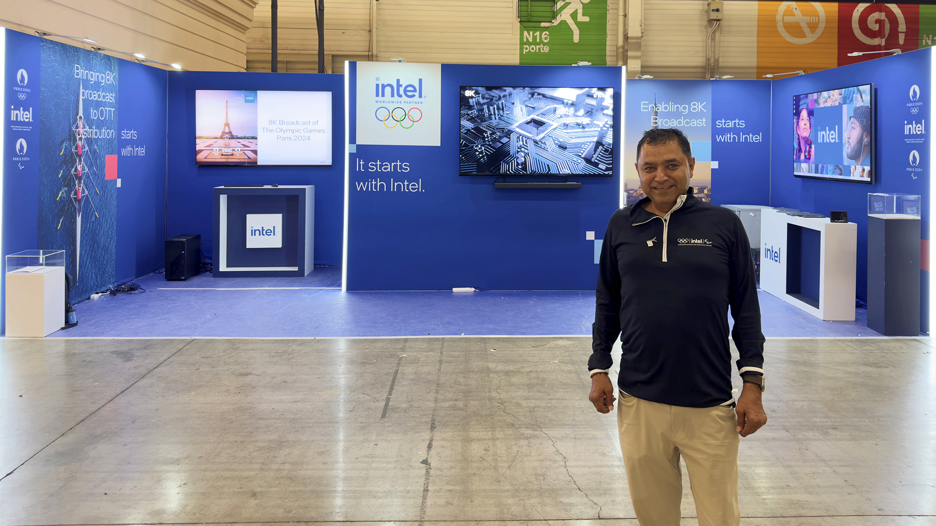 A man stands smiling in front of an Intel exhibition booth at a trade show. The booth features displays with the Intel logo, screens showing technology-related visuals, and promotional text. The flooring and walls are industrial and polished.