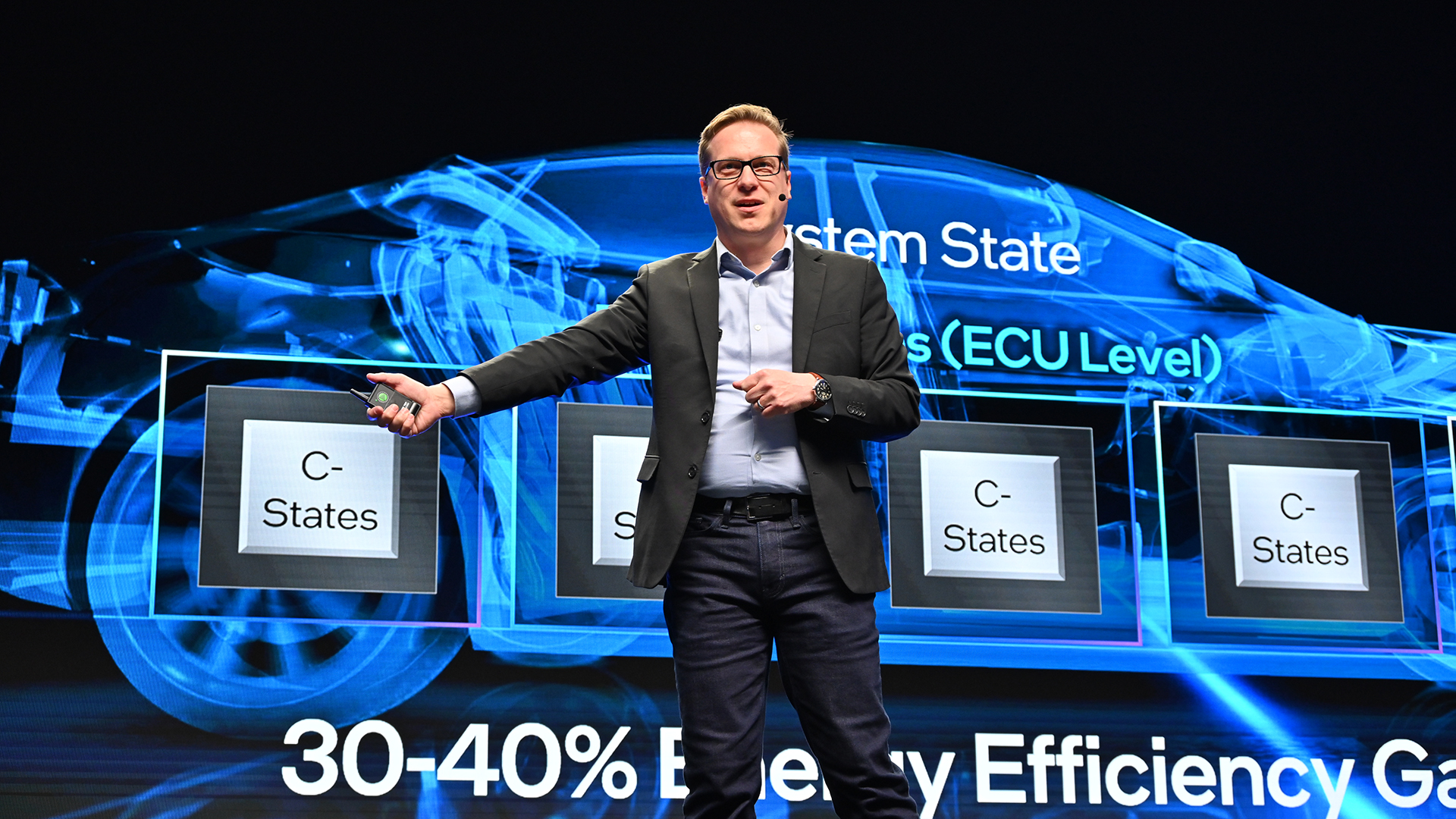 A person in a suit is presenting on stage with a futuristic car design projected behind them. The projection includes text about system states and energy efficiency gains. The person is gesturing with their hand.