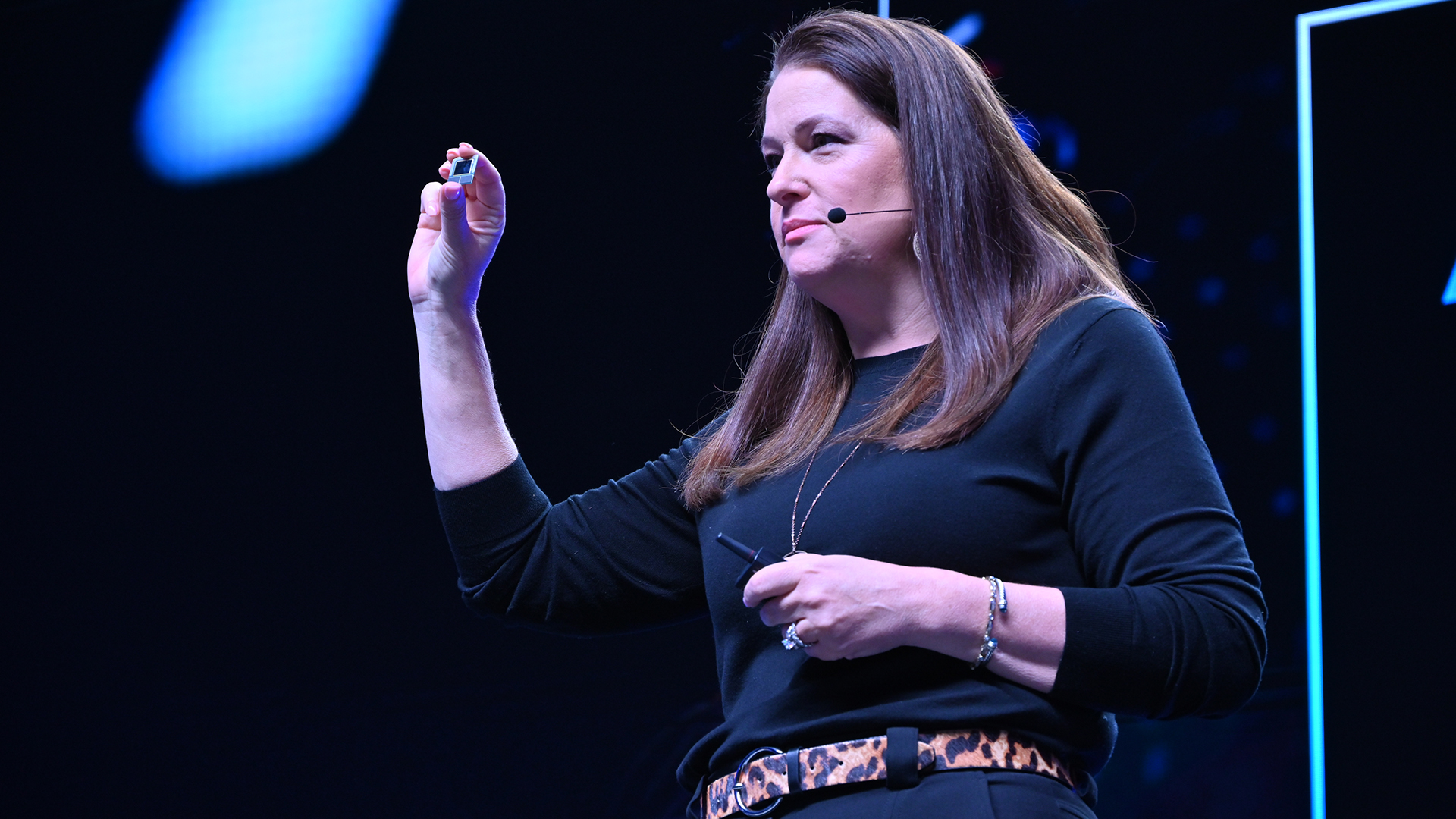 A woman in a black outfit is speaking on stage with a headset microphone. She is gesturing with her hand, holding a small object. The background is dark with blue lighting.