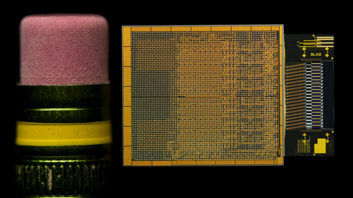 Close-up of a pink pencil eraser next to a small, intricate microchip with a grid of gold circuits on a black background, highlighting the size comparison.