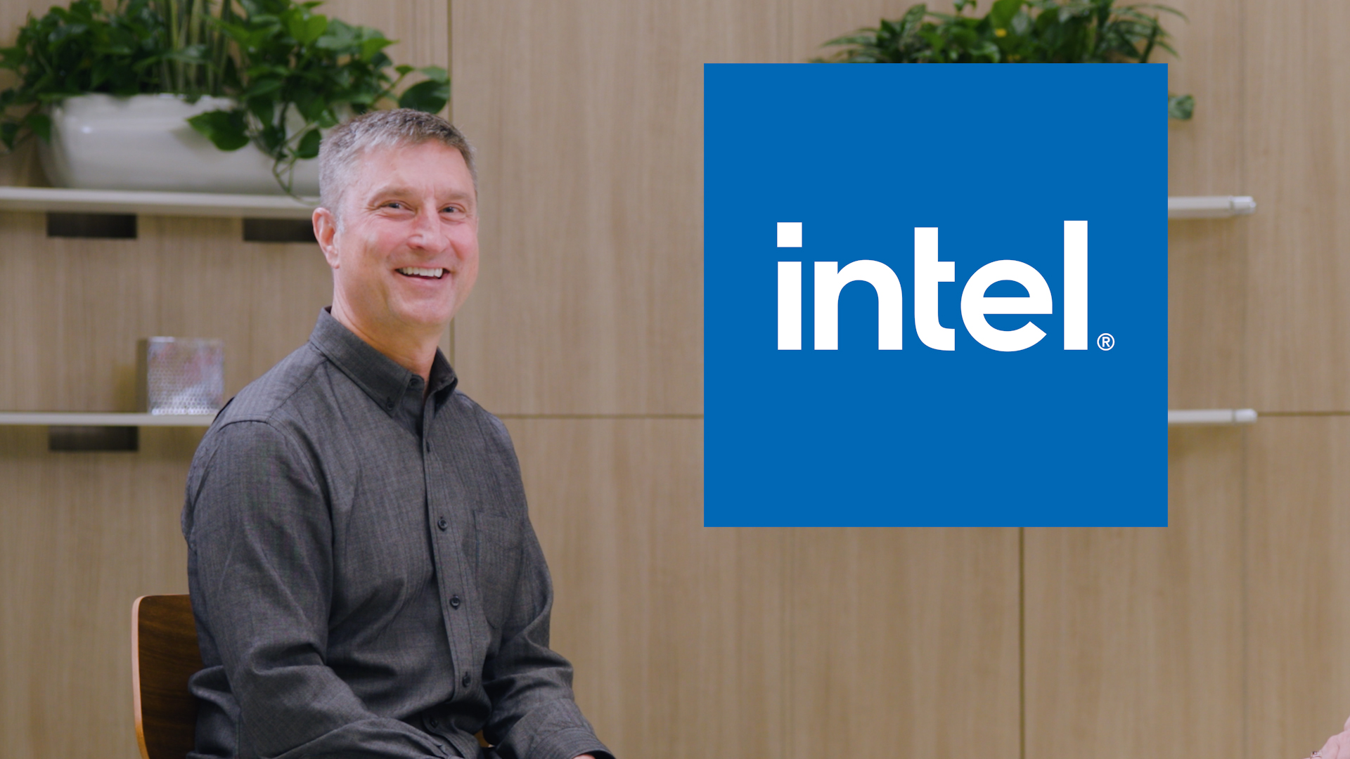 A person sits smiling on a chair in a modern office setting with wooden walls and potted plants. To the right, the blue Intel logo is displayed prominently.