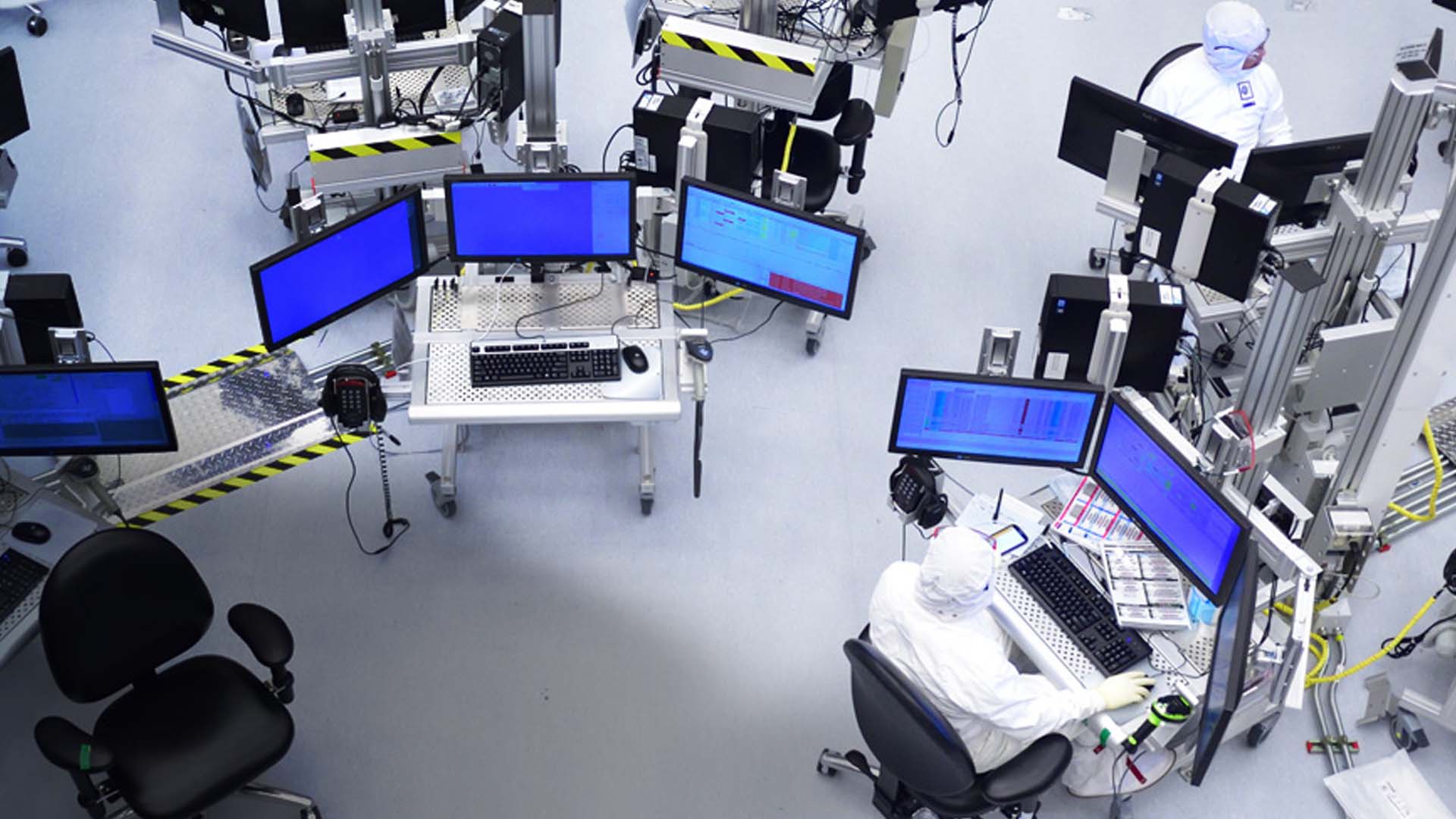 A high-tech laboratory with several workstations equipped with multiple monitors and computers. People in white protective suits are seated, working on the computers, surrounded by industrial equipment and cables on a clean, gray floor.