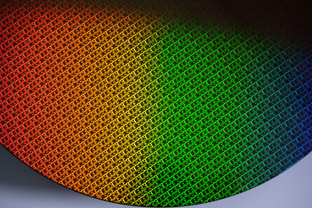 A close-up image of a silicon wafer displaying a spectrum of colors including red, orange, yellow, green, and blue. The surface is covered with intricate microchip patterns, demonstrating a grid-like texture.