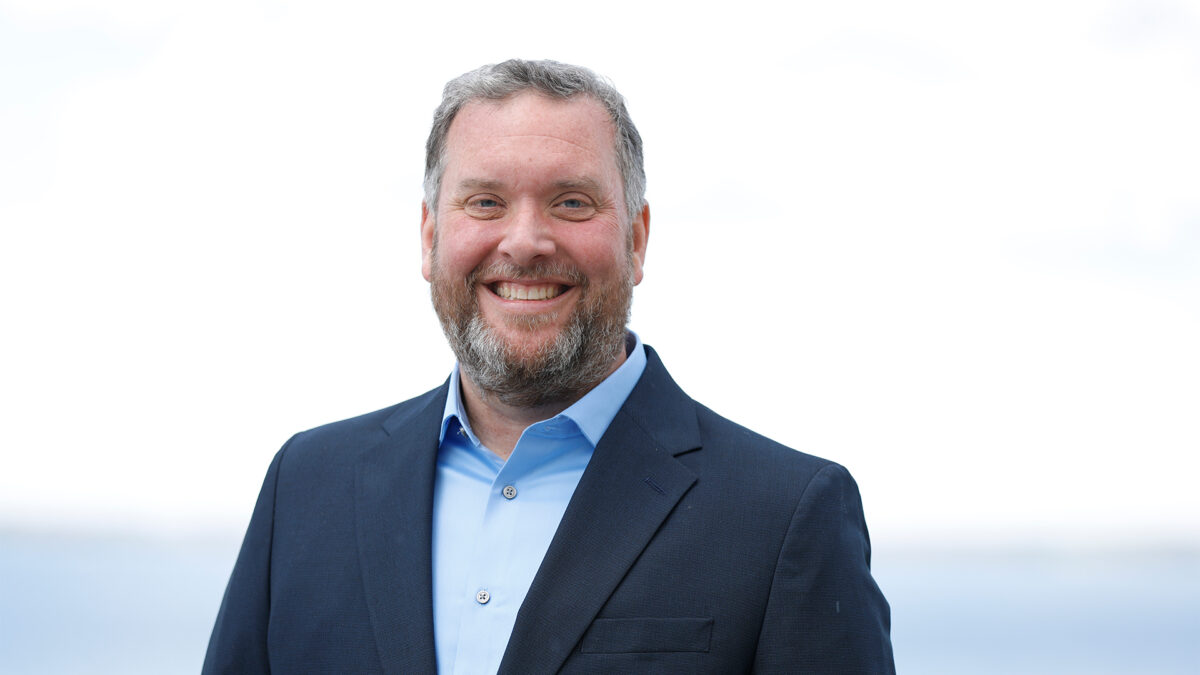 A smiling man with a beard wearing a blue button-up shirt and a dark suit jacket stands outdoors against a bright, blurred background.