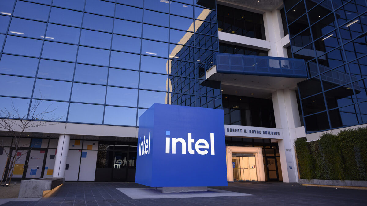 A large blue cube with the Intel logo stands in front of a modern glass office building, labeled Robert N. Noyce Building. The sky and surrounding structures reflect in the glass facade.