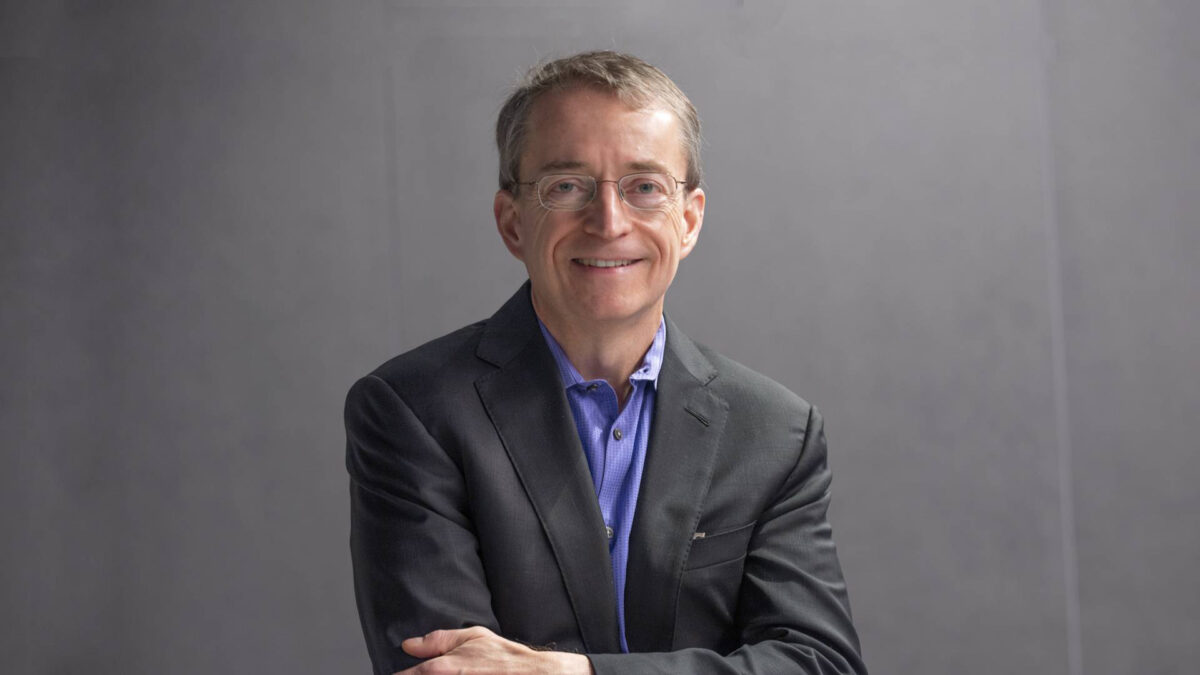 A person wearing glasses, a dark suit, and a blue shirt is smiling with arms crossed against a neutral gray background.