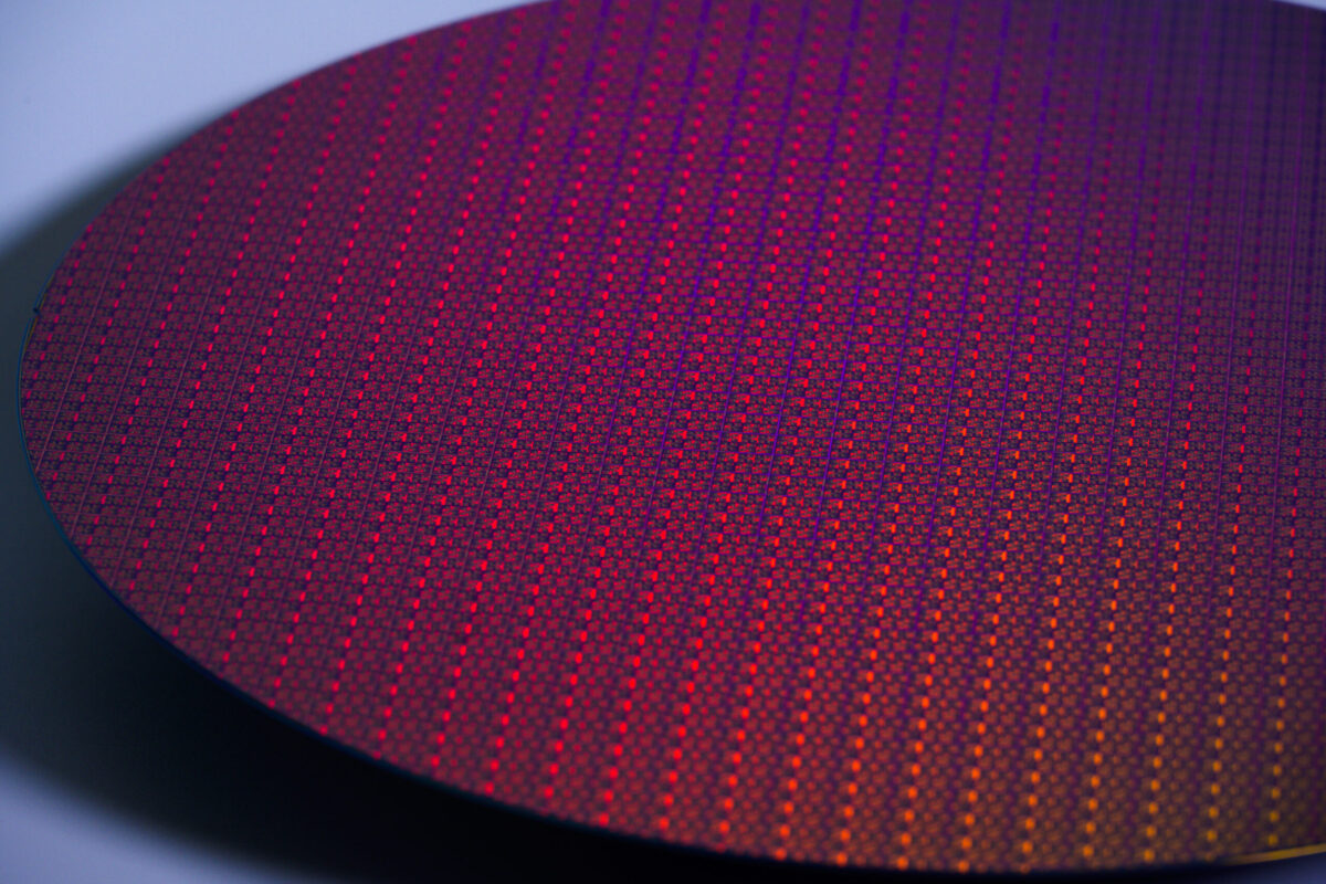 Close-up of a vibrant semiconductor wafer, displaying a grid pattern of tiny, multicolored squares. The surface reflects hues of red, purple, and orange, creating a colorful and intricate mosaic effect under the light.