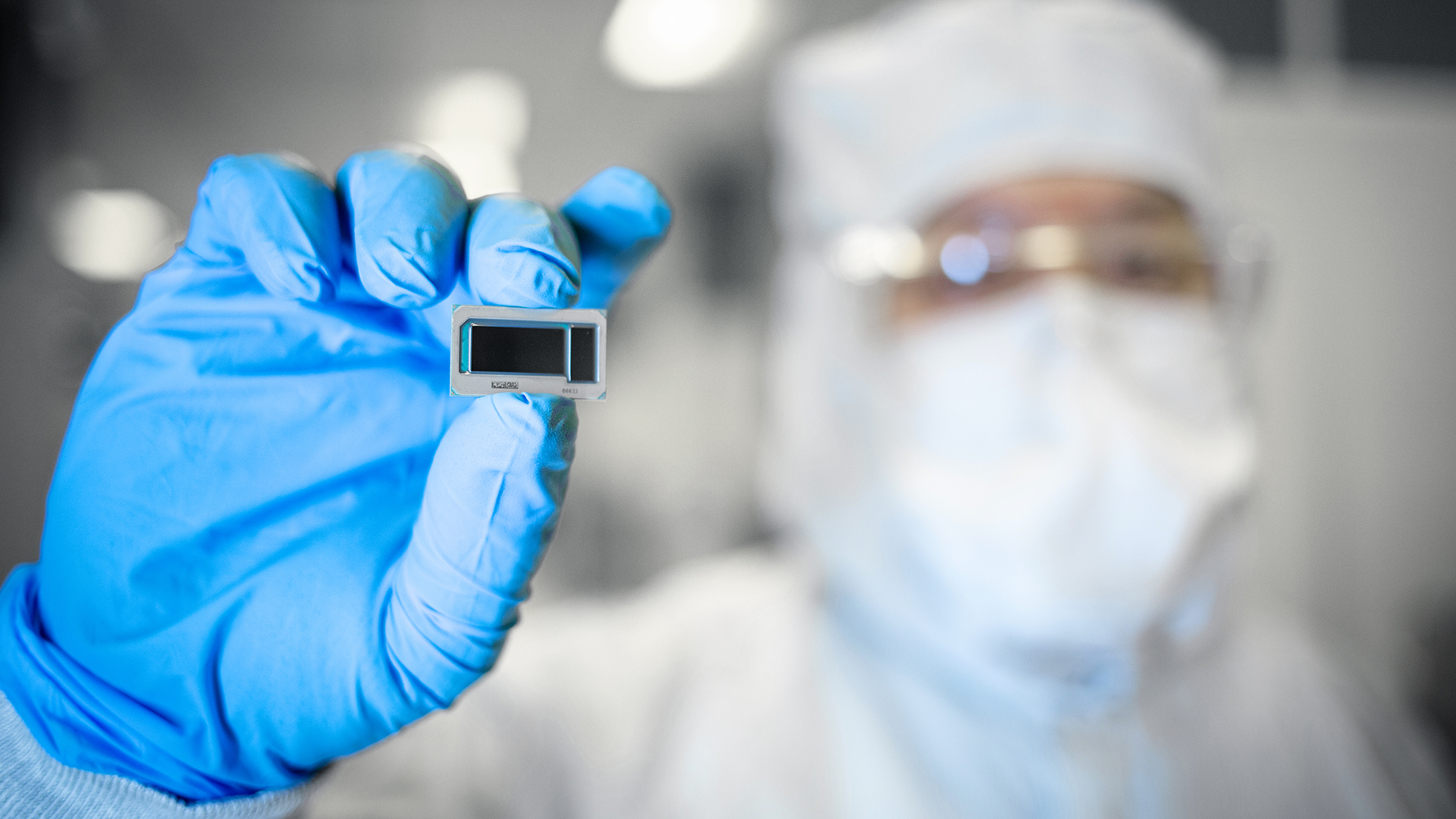 A person wearing protective gear and blue gloves holds a small electronic component, possibly a microchip, between their fingers. The background is blurred, indicating a laboratory or cleanroom environment.