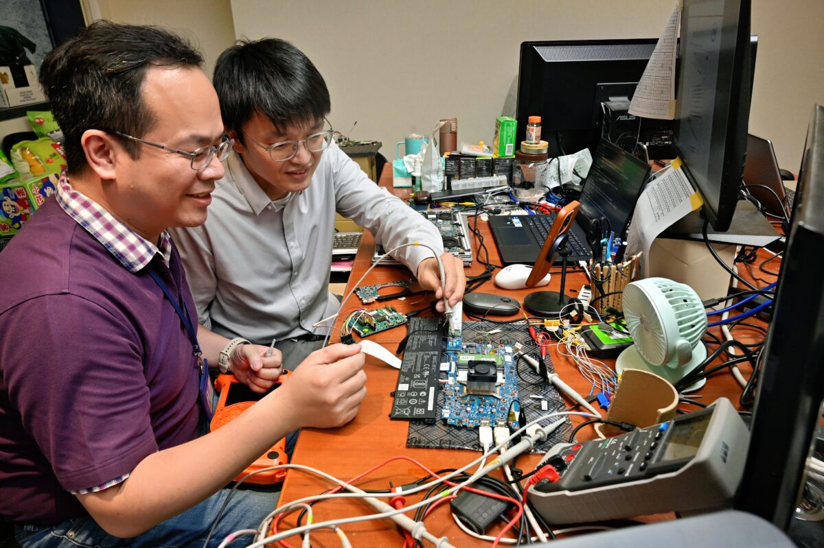 Two people are working on electronic circuits at a cluttered desk filled with various electronic components, tools, and monitors. They appear focused and engaged in their task, surrounded by wires and devices.