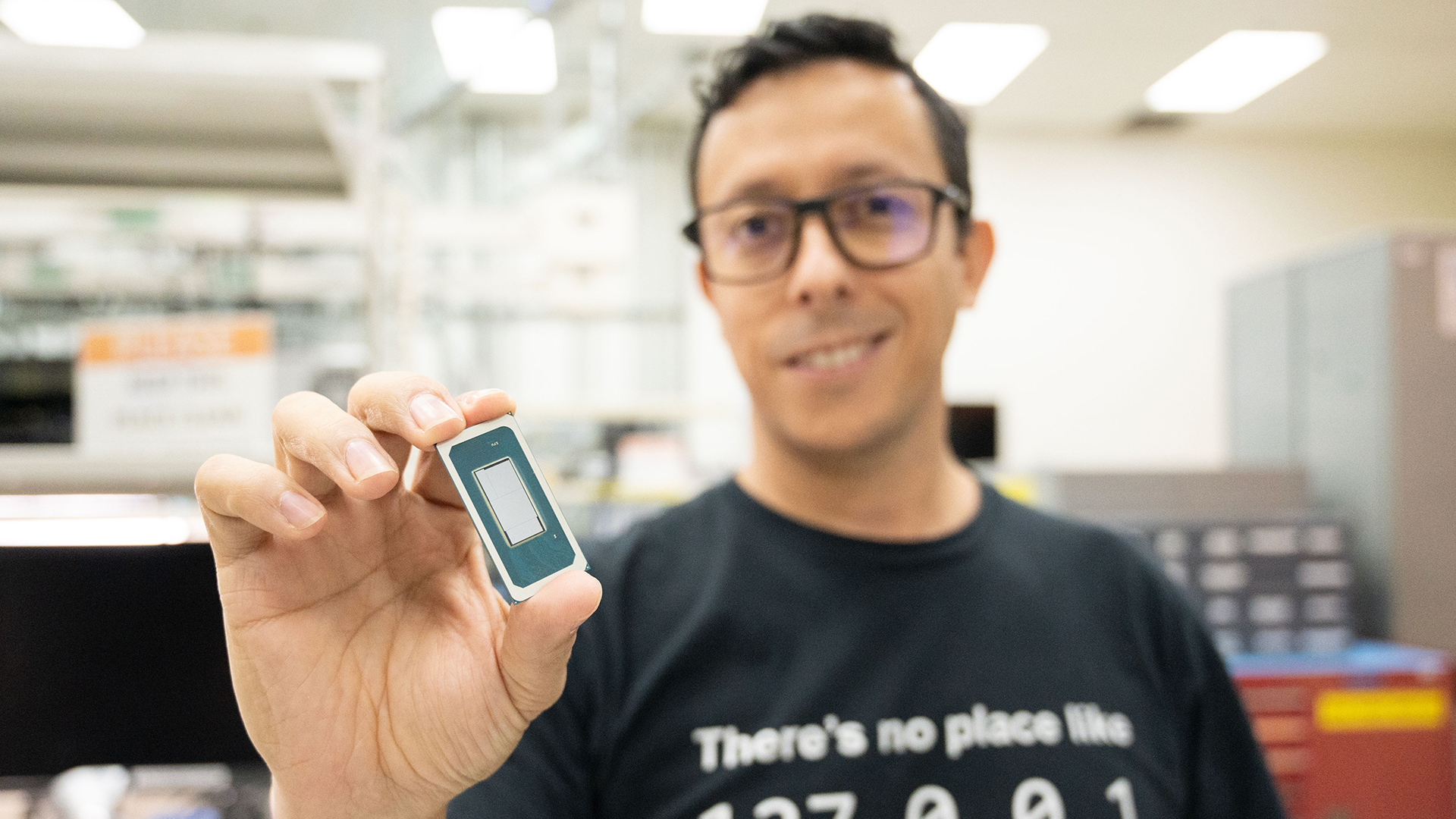 A person wearing glasses and a black t-shirt is holding up a small electronic component in a laboratory setting. The background is blurred, showing shelves and equipment. The t-shirt has a printed message.