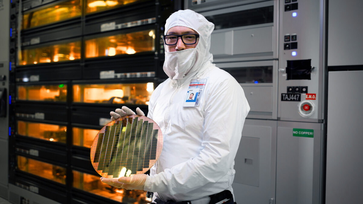 A person wearing a cleanroom suit and glasses holds a round wafer inside a high-tech manufacturing facility. The background shows equipment and illuminated panels.