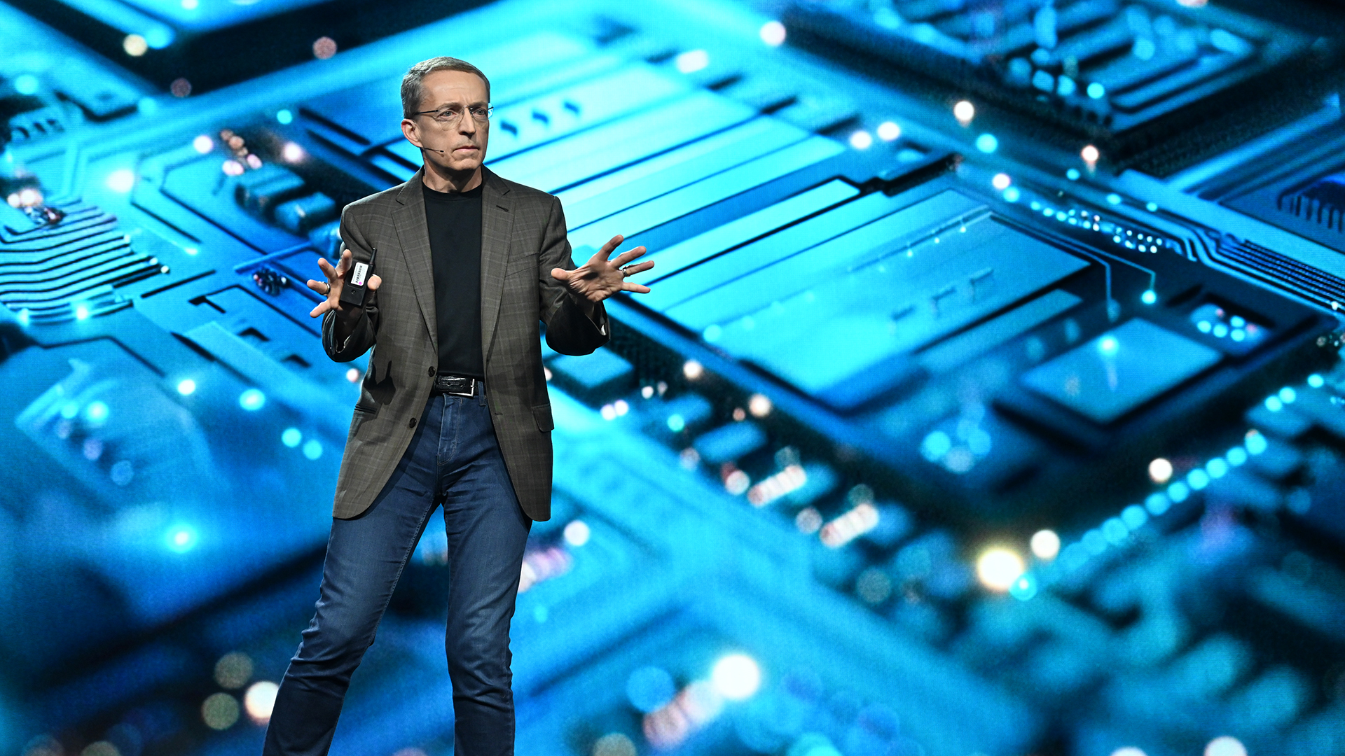 A person in a blazer and jeans speaks on stage with a digital circuit board background, illuminated in blue, creating a tech-themed atmosphere.