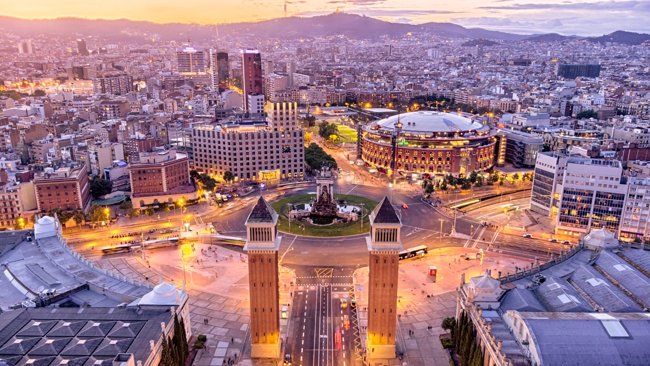 Aerial view of a cityscape at sunset, featuring a large circular building with a lit rooftop. Twin towers frame a wide avenue leading to a central plaza. Buildings and streets are illuminated, with distant hills under a vibrant sky.