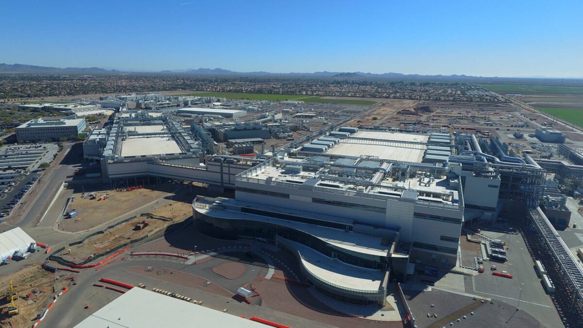 Aerial view of a large industrial complex with multiple white-roofed buildings, surrounded by roads and open land under a clear blue sky. Residential areas and distant mountains can be seen in the background.